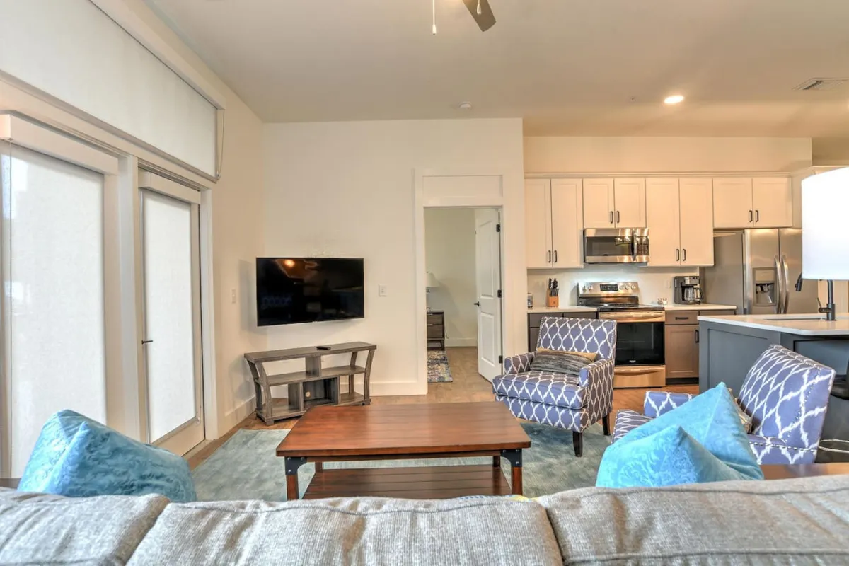 A spacious, light-filled living area featuring a plush sofa and armchairs arranged around a coffee table, with a television and an adjacent kitchen in view. Large windows let in ample natural light, and the open layout invites relaxation and socializing.