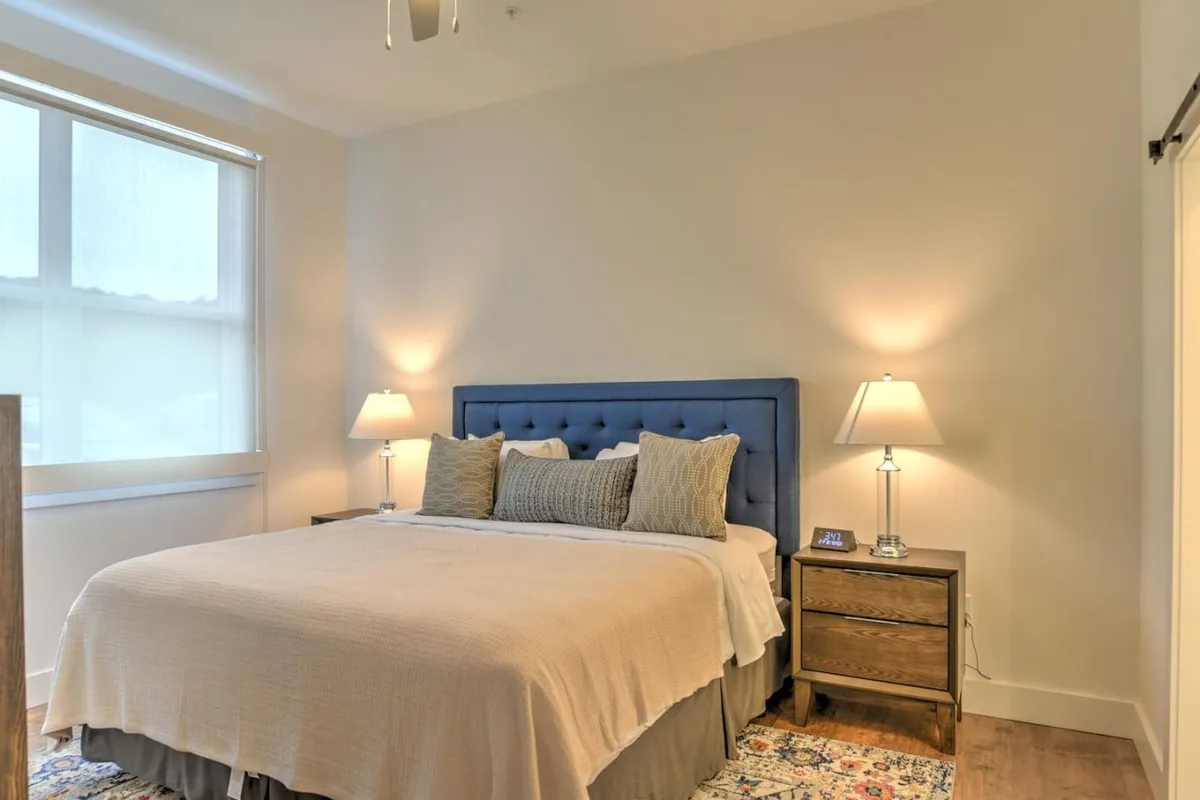 A bright king bedroom with a comfortable bed, nightstands topped with table lamps, a cozy rug underfoot, and a large window filling the room with natural light. The minimalist, soothing décor invites restful sleep and relaxation.