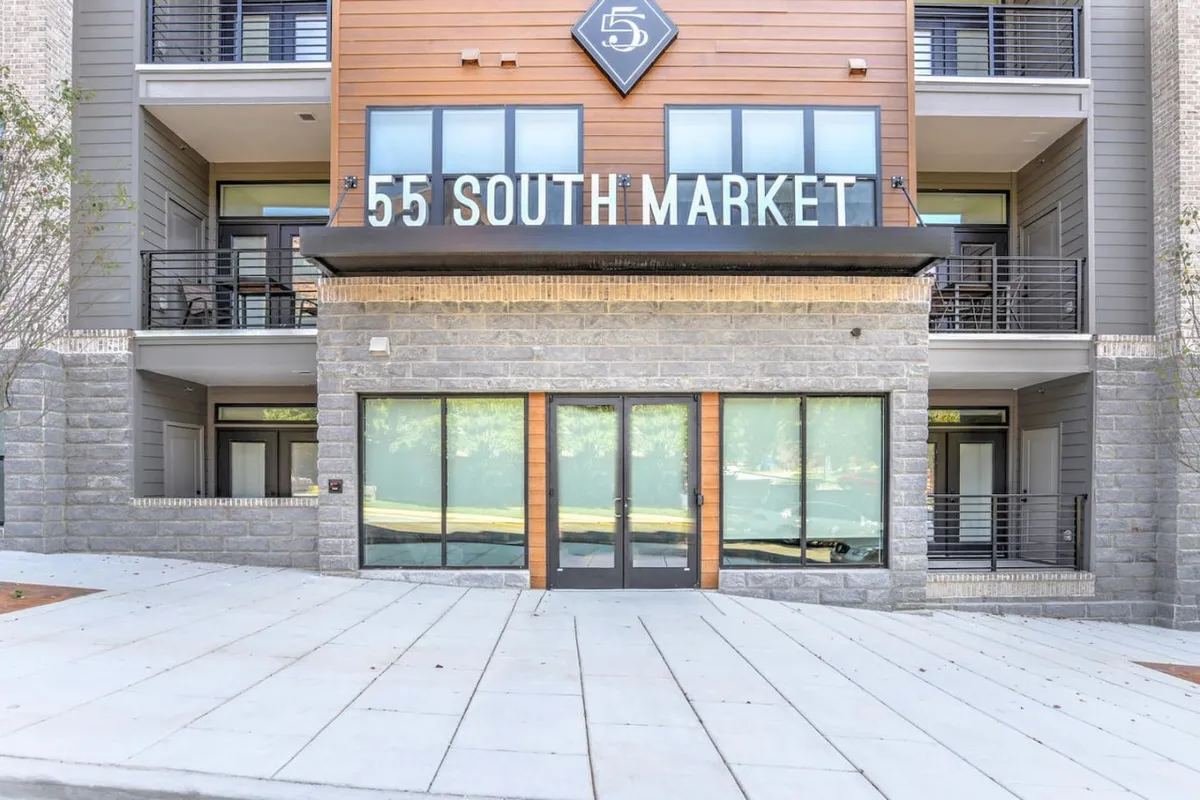 This image captures the exterior of the building with the '55 SOUTH MARKET' sign, a wooden and stone facade, and expansive glass windows. The inviting exterior hints at access to private balconies above, with the building set against a backdrop that could suggest city views.
