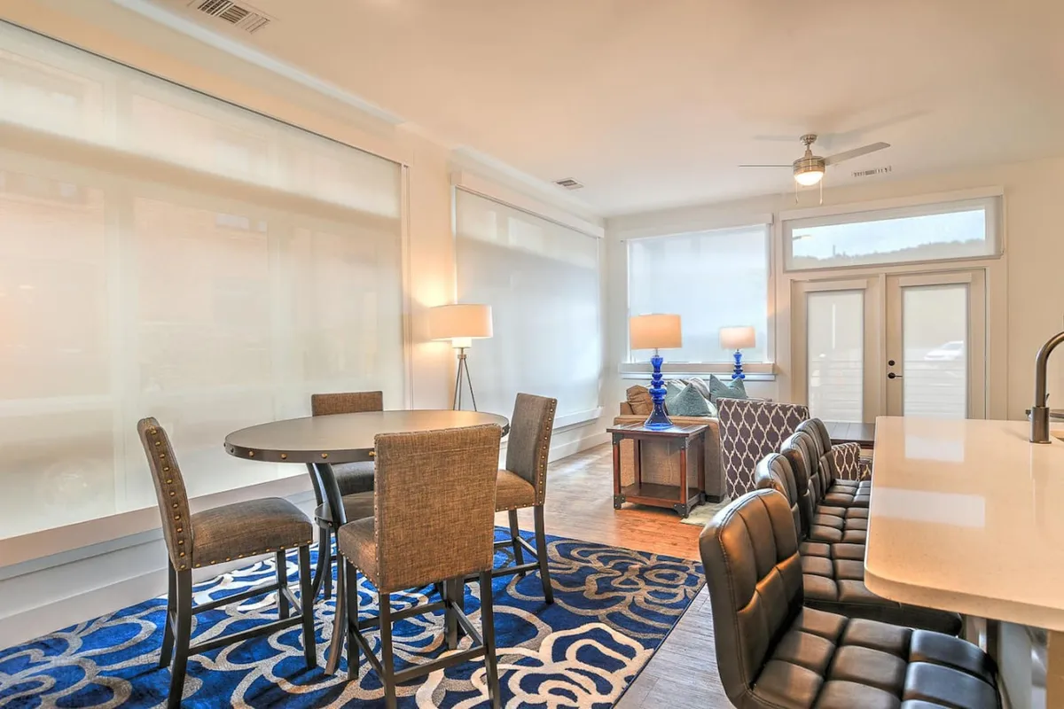 A welcoming open-concept living and dining area with a stylish blue area rug, a well-set dining table, and comfortable seating. Large windows suggest access to the outdoors and a vibrant urban environment beyond.