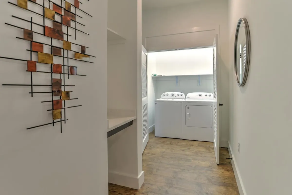 A tidy laundry area with a modern washing machine, decorative wall art, and a large mirror, conveying convenience and practical amenities for longer stays.