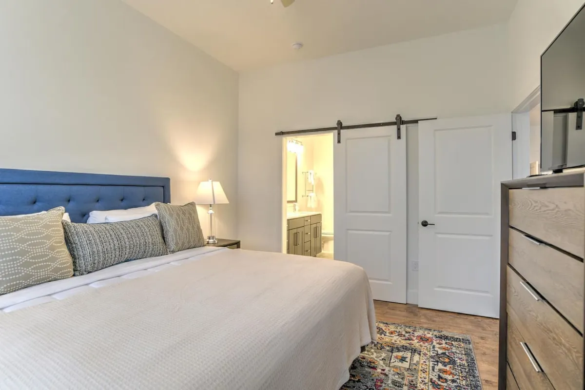 A tranquil king bedroom featuring a plush king-sized bed with a blue headboard, layered decorative pillows, a nightstand with a lamp, a dresser, and a sliding barn door leading directly to an ensuite bathroom. The calming tones and tasteful décor create a restorative, private retreat.