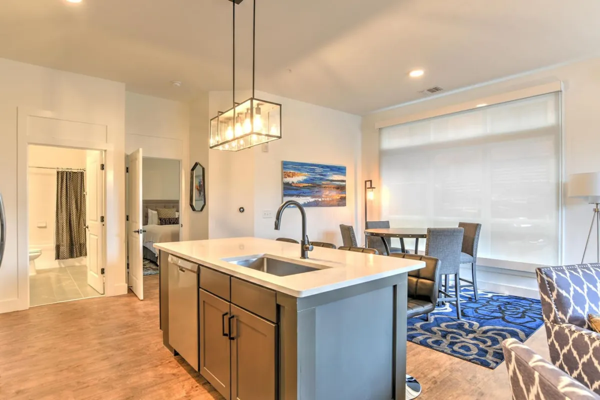 The image centers on a bright, modern kitchen featuring a generous kitchen island, stainless-steel appliances, and an adjacent dining table. The open layout invites connection between the kitchen and seating areas, with bold artwork adding a contemporary flair. The space is staged for cooking and dining, reflecting a welcoming, social atmosphere.